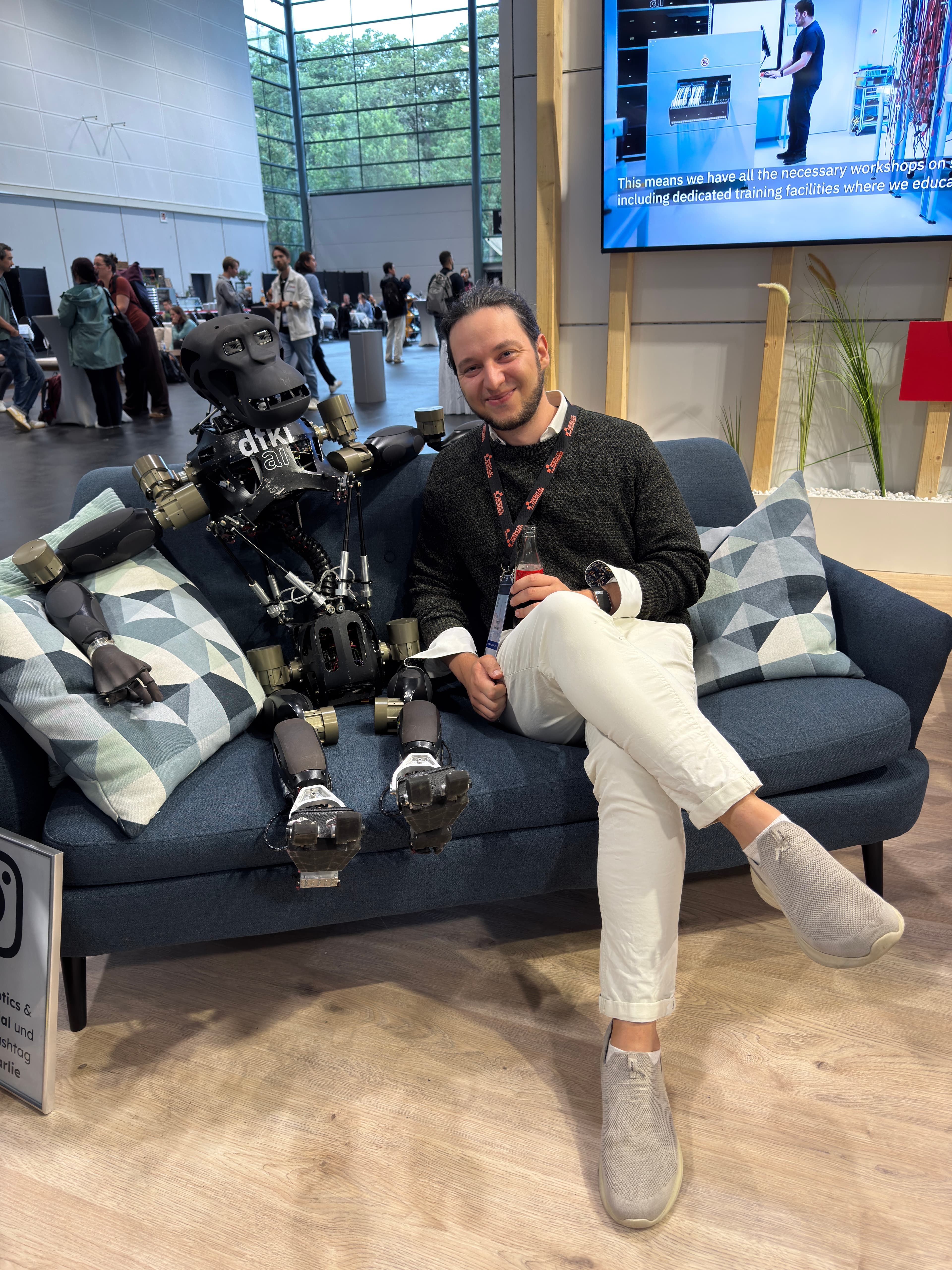 Georg Roch sitzt lächelnd neben einem humanoiden Roboter auf einem Sofa.
